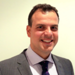 A headshot image of Mr Panos Dimitriadis. He is photographed smiling toward the camera wearing a grey suit and tie.