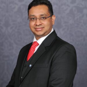 A professional headshot photograph of Dr Ritesh Kumar. He is pictured wearing a black suit with red tie, smiling toward the camera.